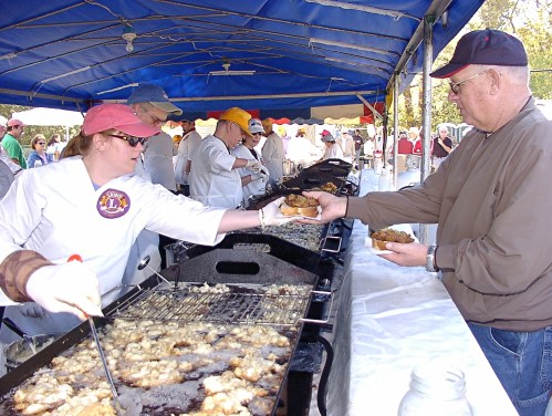 A vendor hands a plate of food to a customer at an outdoor food stall under a blue tent.