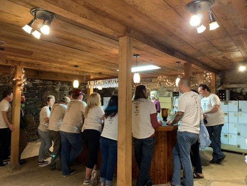 A group of people in matching shirts gather around a wooden bar in a rustic setting, enjoying drinks and conversation.