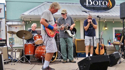 A band performs outdoors, featuring a guitarist, drummer, bassist, and harmonica player, with instruments and amplifiers visible.