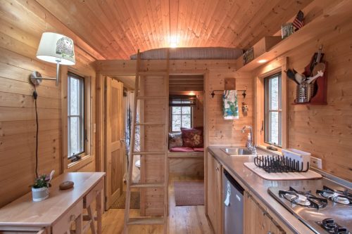 Cozy wooden interior of a tiny cabin featuring a kitchen, ladder, and seating area with natural light.
