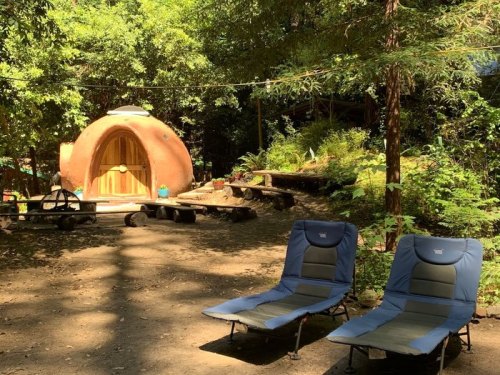 A cozy, round earthen structure surrounded by trees, with two blue lounge chairs in the foreground.