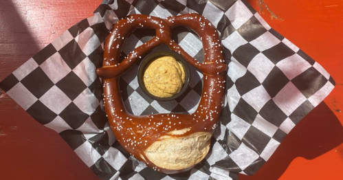 A large, soft pretzel with salt, served in a basket with a small cup of mustard on a checkered paper.