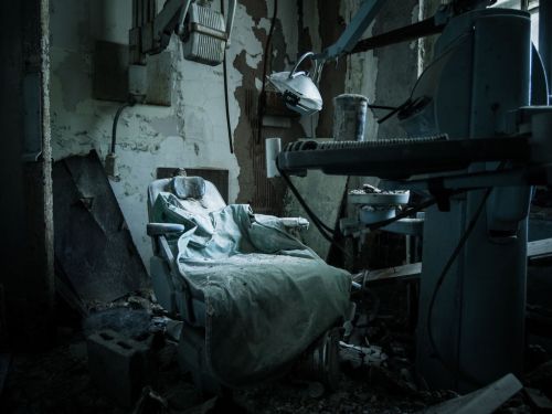 An abandoned dental chair covered in dust, surrounded by debris in a dilapidated room.