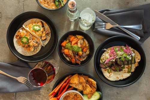 A spread of colorful dishes including tacos, roasted vegetables, and chips on a table with drinks and utensils.