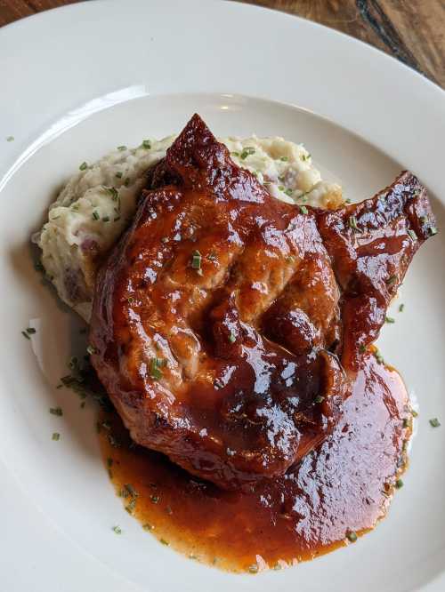 A juicy pork chop glazed with sauce, served on a bed of creamy mashed potatoes, garnished with chives.