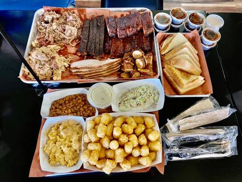 A large spread of barbecue meats, sides like mac and cheese and coleslaw, and sauces on a tray and plates.