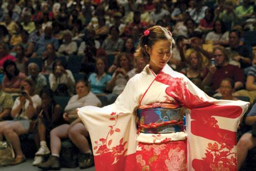 A woman in a colorful kimono performs on stage, with a diverse audience watching attentively in the background.