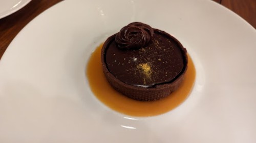 A chocolate dessert with a glossy top, decorated with a chocolate swirl, on a white plate with a caramel sauce.