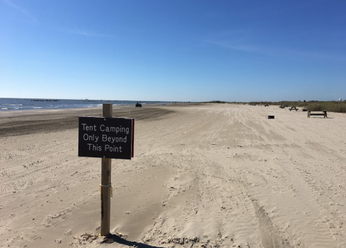 A sandy beach with a sign indicating tent camping is allowed beyond that point, under a clear blue sky.