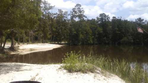 A serene lakeside scene with sandy shore, lush trees, and an American flag gently waving in the breeze.