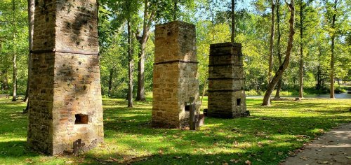 Three stone chimneys stand in a green park surrounded by trees, with a calm water body visible in the background.