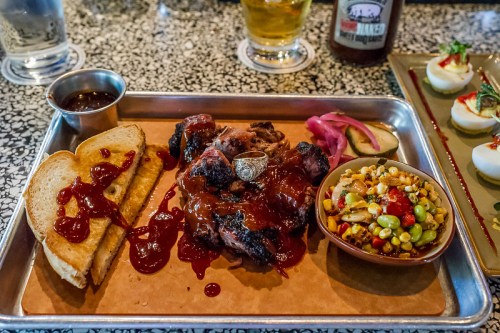 A metal tray with BBQ meat, toast, pickled onions, corn salad, and sauce, set on a speckled table with a drink in the background.