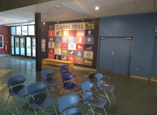 Interior of Slater Mill with blue chairs arranged in front of a colorful wall display featuring various icons and symbols.