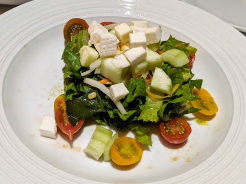 A fresh salad with mixed greens, cherry tomatoes, cucumber, and cubes of feta cheese on a white plate.