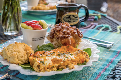 A plate with a vegetable omelet, biscuit, fruit cup, and a cinnamon roll, set on a colorful tablecloth.