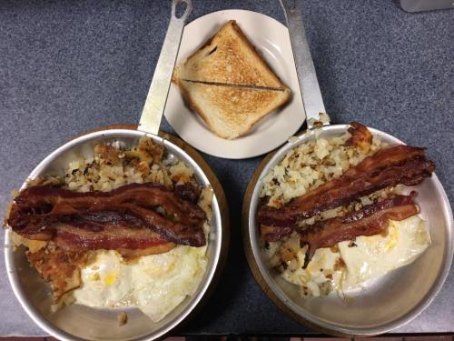 Two small pans with eggs, bacon, and hash browns, alongside a plate with a toasted sandwich.