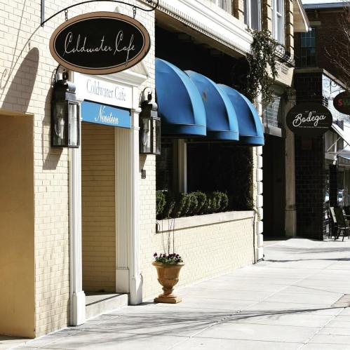 A sunny street scene featuring the entrance of Coldwater Cafe with blue awnings and nearby shops.