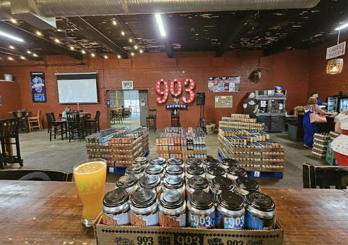 A brewery interior featuring shelves of canned beer, a large "903" sign, and a glass of beer on the bar.