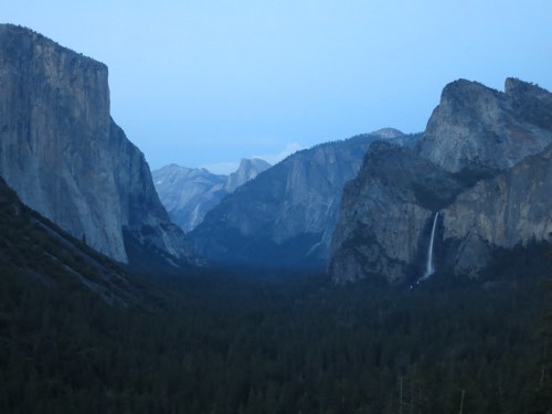 A serene view of Yosemite Valley, featuring towering cliffs and a waterfall surrounded by lush forest at dusk.