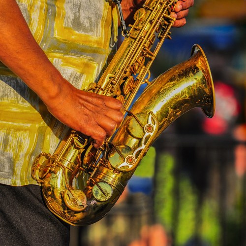 A close-up of a person's hand playing a shiny saxophone, wearing a patterned yellow shirt.