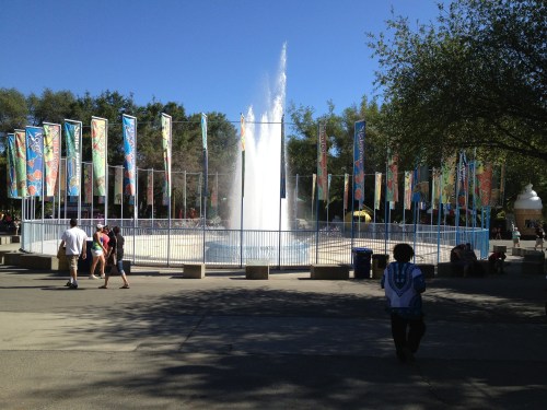 A fountain surrounded by colorful banners, with people walking nearby on a sunny day.