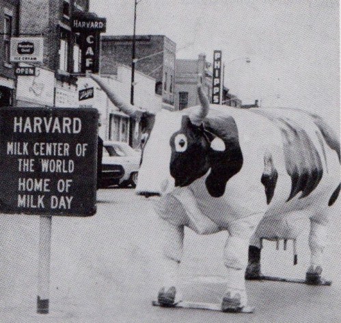 A large inflatable cow stands near a sign reading "Harvard Milk Center of the World, Home of Milk Day" on a city street.
