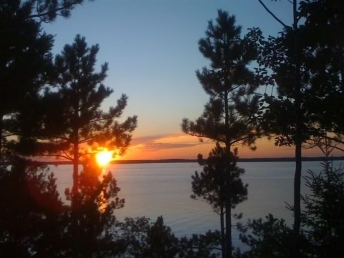 Sunset over a calm lake, framed by silhouettes of pine trees against a colorful sky.