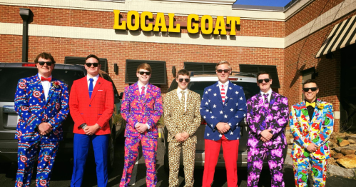 A group of seven men in colorful, patterned suits stand outside a restaurant called "Local Goat."