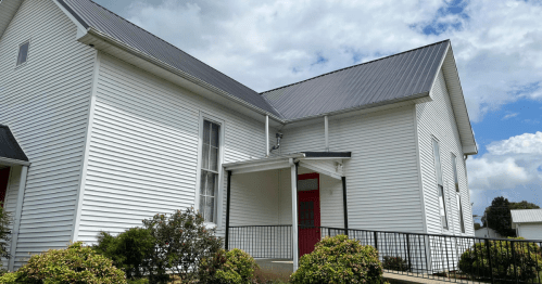 A white building with a metal roof, featuring a red door and surrounded by greenery under a cloudy sky.