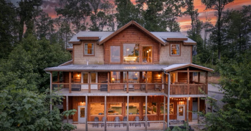 A large wooden cabin surrounded by trees, featuring multiple levels and a porch, set against a colorful sunset sky.