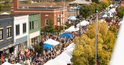 A bustling street fair with crowds, vendor tents, and colorful autumn trees lining the street.