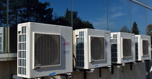 Row of air conditioning units mounted on a wall, reflecting trees and blue sky in nearby glass.