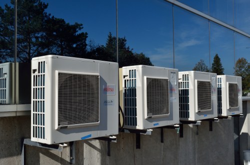 Four air conditioning units mounted on a building's exterior, reflecting trees and sky in the glass windows.
