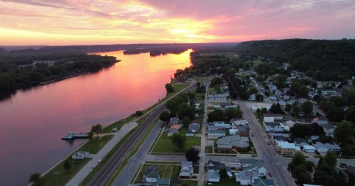 Aerial view of a river at sunset, reflecting vibrant colors, with a small town and green hills in the background.