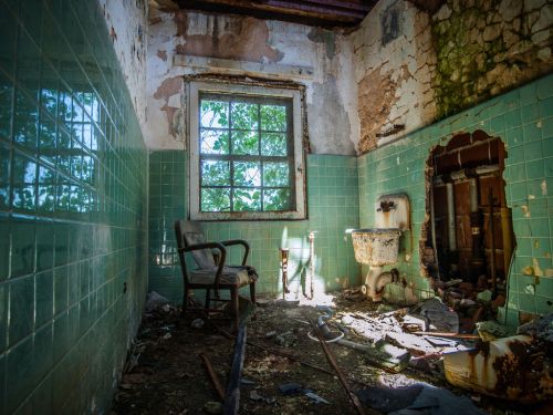 Abandoned bathroom with green tiles, a broken wall, a chair, and sunlight filtering through a window.