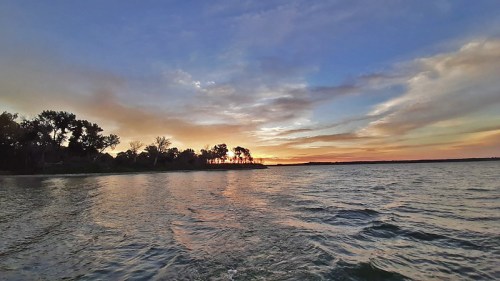 A serene sunset over a calm lake, with silhouettes of trees along the shoreline and colorful clouds in the sky.