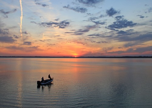 Two fishermen in a boat on calm water at sunset, with vibrant colors and clouds reflecting in the sky.