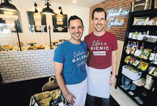 Two smiling men in matching t-shirts stand in a cozy bakery with baked goods displayed behind them.