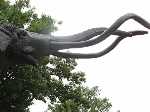A close-up of a large elephant sculpture with long, curved tusks against a cloudy sky and green trees.