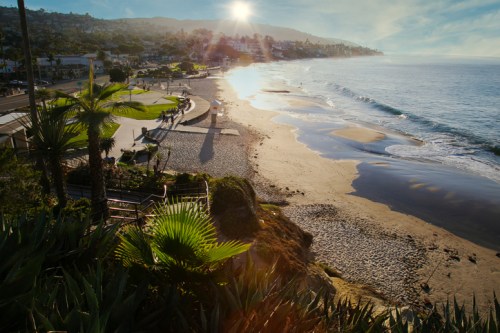 A scenic beach view at sunrise, with gentle waves, palm trees, and people walking along the shore.