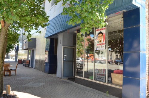A sidewalk view of a storefront with large windows and a sign, surrounded by trees and outdoor seating.