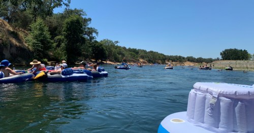 A sunny river scene with people floating on inflatable rafts, surrounded by trees and a clear blue sky.