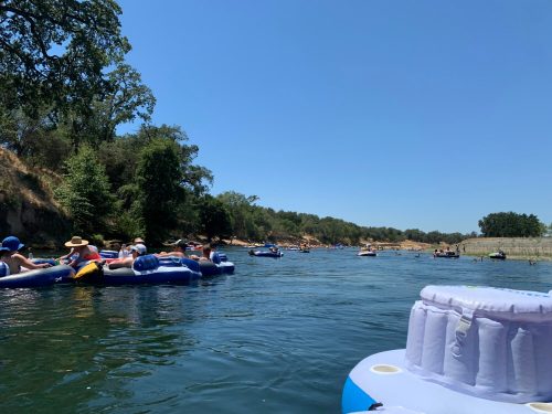 People floating on inflatable rafts in a sunny river surrounded by trees and a clear blue sky.