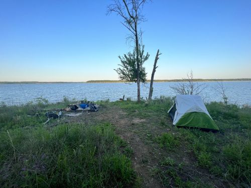 A tent set up by a calm lake, surrounded by greenery and a clear blue sky.