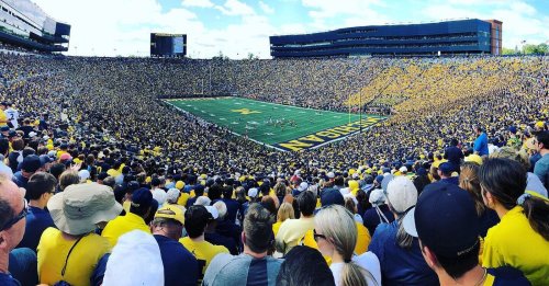 A packed stadium filled with fans in maize and blue, watching a football game on a sunny day.