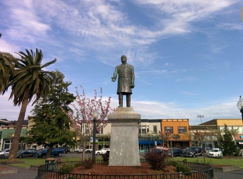 Statue of a man in a coat, surrounded by trees and buildings, under a blue sky with clouds.