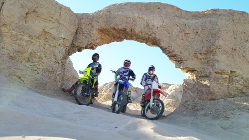 Three motocross riders pose on their bikes under a natural rock arch in a sandy landscape.