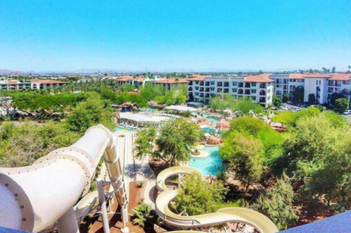 Aerial view of a resort pool area with water slides, surrounded by greenery and buildings under a clear blue sky.