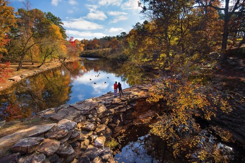A couple stands on a stone bridge by a serene river, surrounded by vibrant autumn foliage and a clear blue sky.