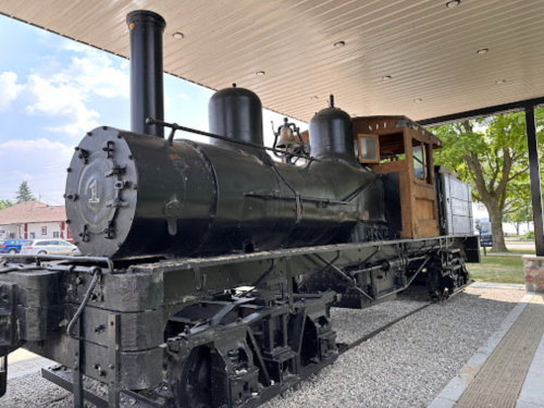 A vintage black steam locomotive displayed under a shelter, with a wooden cabin and surrounded by gravel.